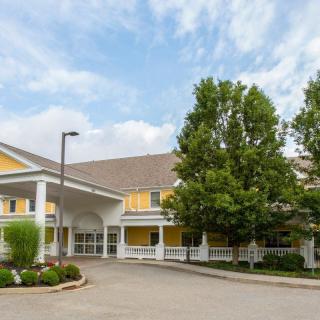 A two-story building with a yellow facade, featuring a covered entrance and surrounded by trees and landscaping under a partly cloudy sky.