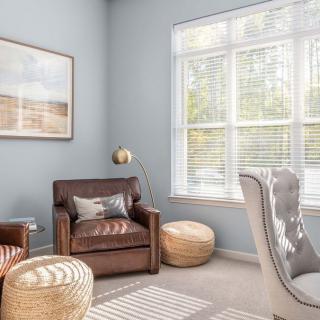 A cozy living room with a leather armchair, poufs, and natural light streaming through the window.