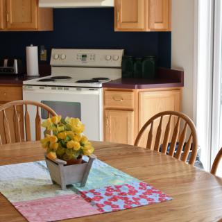 A cozy kitchen with a wooden dining table adorned with a floral centerpiece and surrounded by wooden chairs, with a stove and cabinets in the background.