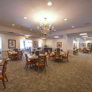 A spacious dining area with neatly arranged tables and chairs under a chandelier, featuring warm lighting and carpeted flooring.