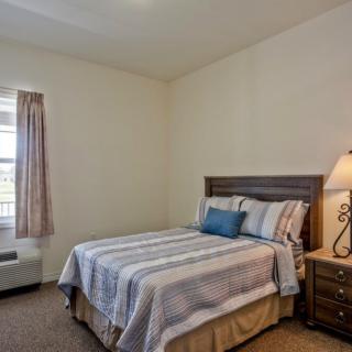 A cozy bedroom with a neatly made bed, nightstand, and a lamp by the window.