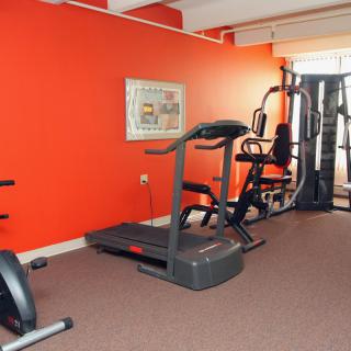 A small gym room with various fitness equipment including a treadmill, exercise bike, and multi-gym machine against a bright orange wall.