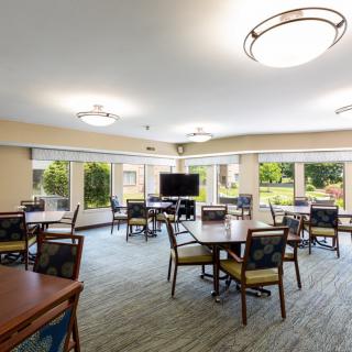 A spacious and well-lit room with multiple tables and chairs arranged for seating, featuring large windows and a television.