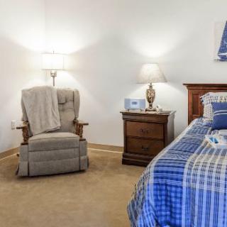 A cozy bedroom featuring a comfortable chair, nightstand with a lamp, and neatly made bed with checkered bedding.