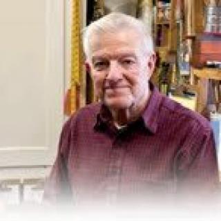 An elderly man sitting indoors with a bookshelf in the background.