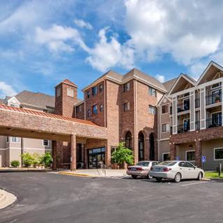 A large brick building with multiple stories, featuring a covered entrance and parking area with cars.