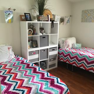 A neatly organized bedroom with twin beds featuring colorful zigzag bedding and a central shelving unit for storage and decor.