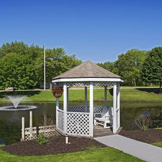 A scenic view of a gazebo by a pond with a fountain, surrounded by lush greenery.