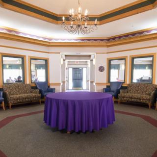 A spacious lobby with a central table draped in a purple tablecloth, surrounded by ornate furniture and illuminated by a chandelier.