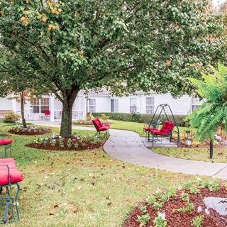 A serene garden setting with a pathway, surrounded by trees and equipped with red-cushioned patio furniture.