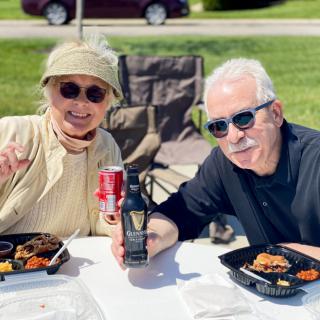An elderly couple enjoying a sunny outdoor picnic with food and drinks.