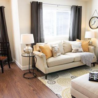 A cozy living room with a beige sofa adorned with mustard and cream throw pillows, flanked by lamps and dark curtains framing a bright window.