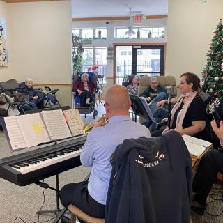 A group of musicians performing indoors with a digital piano, in front of an audience and a Christmas tree.