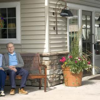 Two elderly individuals enjoying a sunny day on a bench outside a residential building.