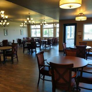 An empty dining area with wooden tables and chairs, illuminated by chandeliers, featuring large windows.