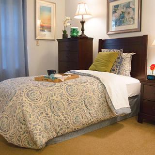 A cozy bedroom with a neatly made bed, complete with patterned bedding and decorative pillows. The room features artwork on the walls, a bedside lamp, and subtle decor elements.