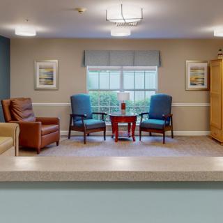 A cozy living space featuring a pair of blue chairs and a brown leather chair arranged around a small wooden table, set in front of a large window with framed artwork on the walls.