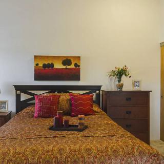 A cozy bedroom with a warm color palette, featuring a bed adorned with patterned bedding and vibrant pillows, complemented by a landscape painting above the headboard. A wooden dresser with a flower vase adds charm to the space.
