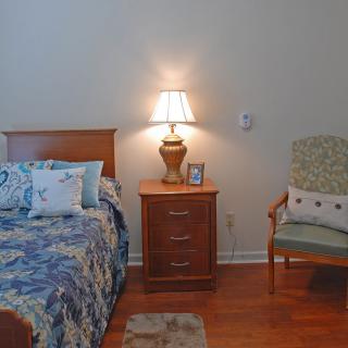 A cozy bedroom with a bed, nightstand, and chair, softly lit by a table lamp.