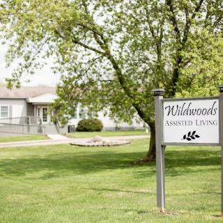 A sign for Wildwood Assisted Living stands in front of a building surrounded by trees and a well-maintained lawn.