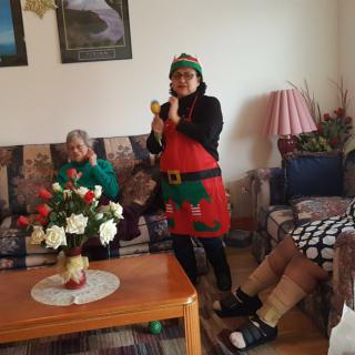 A person dressed in a festive elf costume stands in a living room, with family members seated nearby.