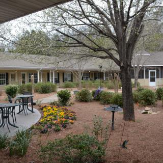A peaceful garden with a patio featuring tables and chairs, surrounded by trees and landscaping.