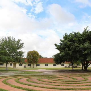 A view of a labyrinth with trees and a building in the background under a partly cloudy sky.