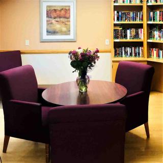 A cozy library reading area with a round table, surrounded by chairs and bookshelves, featuring a vase of flowers as a centerpiece.