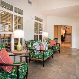 A well-lit lobby area featuring green patterned chairs, colorful cushions, and stylish lamps next to large windows with blinds.