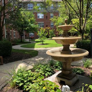 A peaceful courtyard garden with a central fountain surrounded by lush greenery and trees, adjacent to a brick building.