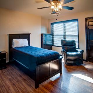 A modern bedroom with a single bed, armchair, and wooden flooring, illuminated by natural light from a large window.