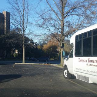 A white bus parked in an empty lot on a sunny day, surrounded by trees and a building in the background.