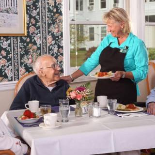 A caregiver serves a meal to an elderly person at a dining table in a warm, welcoming environment.