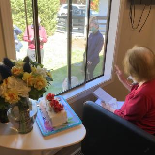 An elderly woman celebrates her birthday indoors with a cake and flowers, while family or friends visit from outside the window.