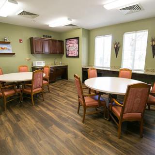 A cozy dining room with round tables and cushioned chairs, featuring wooden flooring and ample natural light from large windows.