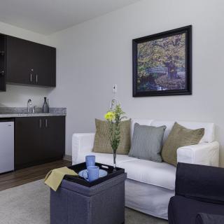 A modern living room with a white sofa, cushions, and a coffee table, adjacent to a kitchenette.