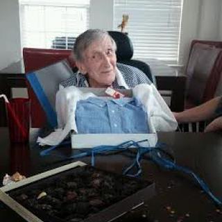 An elderly person sitting in a wheelchair at a table with an open box of chocolates nearby.