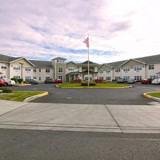 A residential building with a flagpole in front, surrounded by a parking lot and landscaping under a cloudy sky.