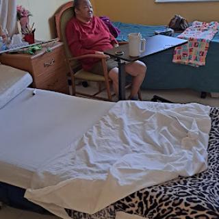 An elderly person sitting in a room with a bed and a table, surrounded by personal items.