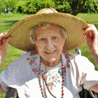 An elderly woman wearing a straw hat and smiling outdoors in a garden on a sunny day.