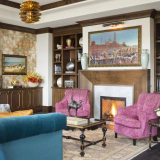 A cozy living room with a fireplace, surrounded by bookshelves and decorated with colorful armchairs and artwork.