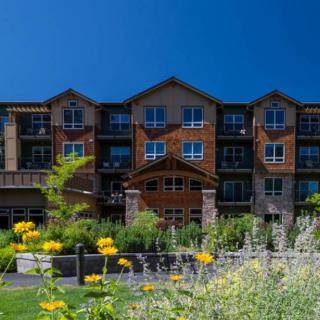 A modern apartment building set against a clear blue sky, surrounded by a landscaped garden with blooming flowers.