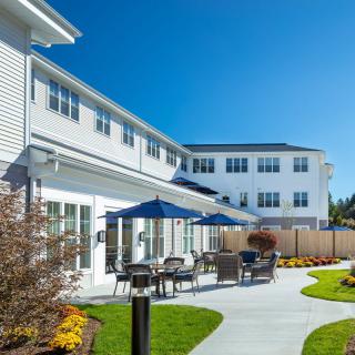A modern building with a sunlit patio area featuring tables, chairs, and umbrellas.