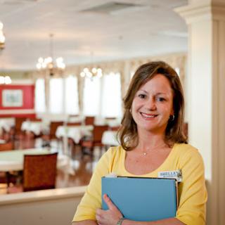 A smiling woman in a yellow shirt holds a folder, standing in a well-decorated restaurant.