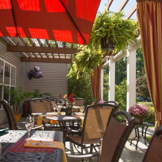 An inviting outdoor patio with tables and chairs under a red umbrella, surrounded by greenery.