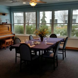 A cozy dining room with a table set for a meal, adjacent to a piano and large windows providing natural light.