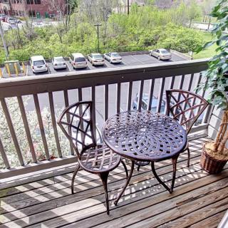 A cozy balcony with a metal table and chairs overlooking a parking lot and greenery.
