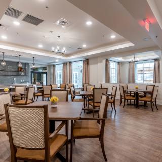 A spacious dining area with wooden flooring, elegantly arranged tables and chairs, and ample natural light from large windows.