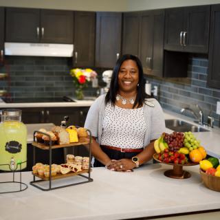 A woman standing in a kitchen surrounded by a display of fruit, baked goods, and lemonade.