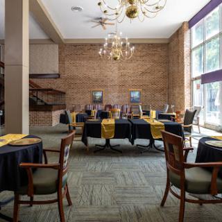 An elegantly arranged dining area featuring tables covered with black cloths and yellow napkins, complemented by a bright ambiance from large windows and decorative chandeliers.